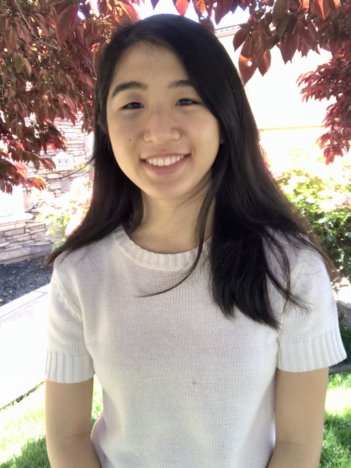 A young asian woman with mid-length dark hair wears a light pink t-shirt and smiles at the camera in front of a crimson-leafed tree.